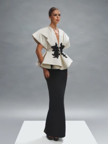 Вечернее платье couture collection 2026 года выполнено в современном, архитектурном и элегантном стиле, представленное длинным прямым силуэтом с архитектурным пеплумом, создающим объем в области талии. Структурированный лиф с глубоким V-образным вырезом дополнен короткими, широкими рукавами-крылышками. Верхняя часть платья выполнена из плотного, хорошо держащего форму дюшес-атласа с легким глянцевым блеском, в цвете слоновой кости, тогда как длинная юбка выполнена из матового, гладкого, струящегося черного крепа. По центру лифа, в области талии, расположены объемные черные аппликации с растительным мотивом.