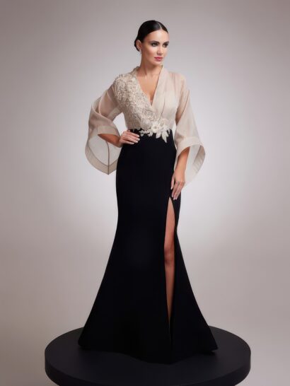 Вечернее платье 23594 couture collection — длинное, современное и элегантное изделие в силуэте годе, облегающем фигуру до коленей и плавно расширяющемся к низу, с умеренным шлейфом. Юбка дополнена высоким боковым разрезом по левой ноге. Лиф с запа́хом формирует глубокий V-образный вырез. Одна сторона лифа выполнена из полупрозрачной ткани, другая — из плотной основы с плотной объемной вышивкой бисером и пайетками в тон, расположенной на одном плече и в верхней части груди. Линию талии подчеркивают объемные цветочные аппликации в белых и молочных тонах. Верх платья дополняют объемные широкие рукава кроя «летучая мышь», выполненные из полупрозрачной ткани и достигающие запястья. Спинка оформлена высоким воротником-стойкой и вертикальным разрезом от основания шеи до поясницы. В верхней части разрез образует вырез типа «замочная скважина», частично закрытый рядом мелких пуговиц. Верхняя часть спинки, как и рукава, выполнена из полупрозрачной ткани. Застежка состоит из ряда мелких пуговиц вдоль верхнего края вертикального разреза спинки и потайной молнии по центральному шву черной юбки, спускающейся от талии. Верх платья, включая объемные рукава и полупрозрачную сторону лифа и спинки, выполнен из легкого, струящегося полупрозрачного шифона светло-бежевого (нюдового) оттенка, создающего воздушный объем. Контрастная сторона лифа и область вышивки используют плотную, матовую ткань с рельефной текстурой. Облегающая юбка-годе изготовлена из плотного матового черного крепа с гладкой поверхностью, обеспечивающего структурированный силуэт. Декоративные элементы — вышивка бисером и пайетками, а также цветочные аппликации — выполнены в оттенках бежевого, белого и молочного, гармонично дополняя общую двухцветную палитру.