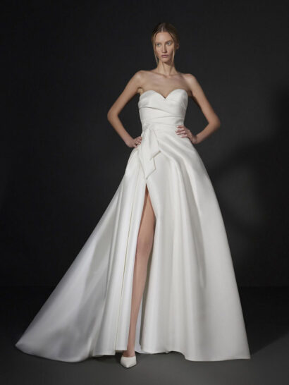 Свадебное платье Vera Wang Jadeau в стиле современного минимализма выполнено из плотного атласа с мягким полуматовым блеском. Структурированный лиф с открытым декольте в форме сердца украшен диагональными драпировками, акцентирующими центр изделия. 



На талии расположен объемный декоративный узел в форме банта, плавно переходящий в глубокие встречные складки пышной юбки, создающие выразительный силуэт и четкие линии. Модель дополнена высоким вертикальным разрезом на юбке и длинным шлейфом «chapel train», подчеркивающим торжественность образа. 



Конструкция платья дополнена скрытой застежкой-молнией, обеспечивающей безупречную посадку. Выверенные пропорции и архитектурный крой подчеркивают баланс между сдержанной элегантностью и современной эстетикой.